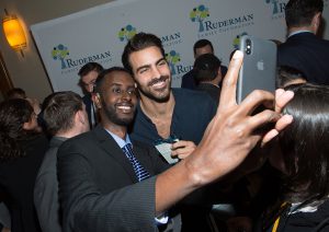 Nyle smiling at a phone next to a fan. A dozen people are milling about in the background. 