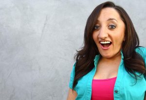 Nicole Evans pictured from the chest up, wearing a bright pink shirt and a bright aqua cardigan, smiling with a surprised look, with a gray wall in the background.